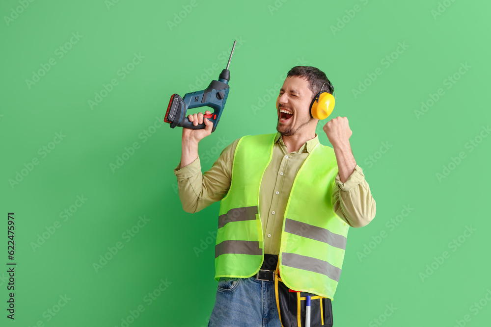 Male builder in hearing protectors with drill on green background