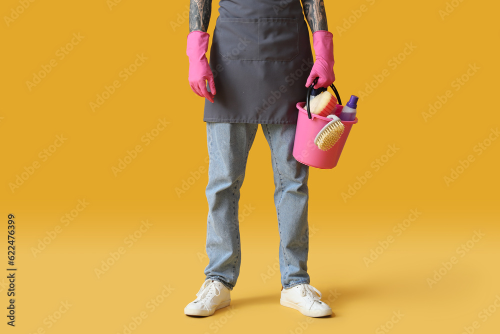 Young tattooed man with bucket of cleaning supplies on orange background