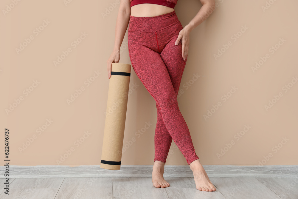 Sporty young woman in leggings and with yoga mat near beige wall