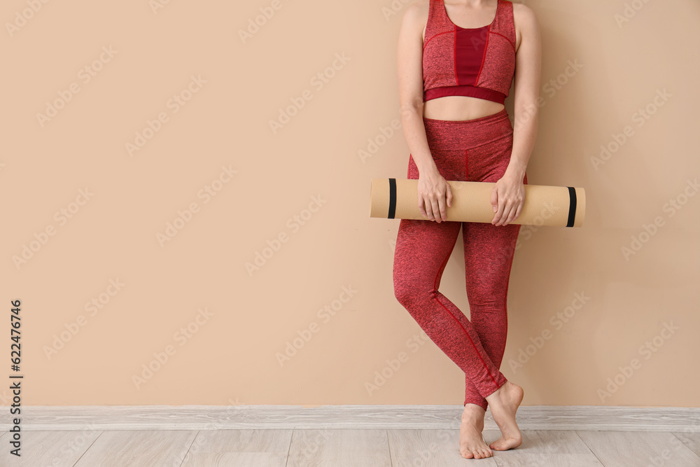 Young woman in sportswear and with yoga mat near beige wall