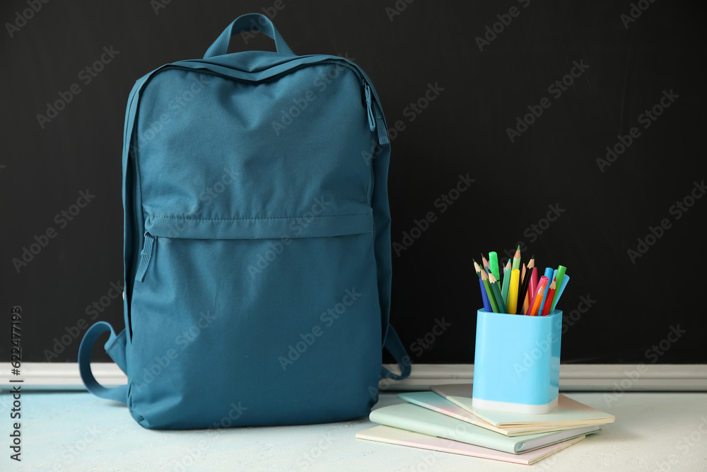 Blue school backpack with notebooks, cup of markers and pencils on white table near black chalkboard