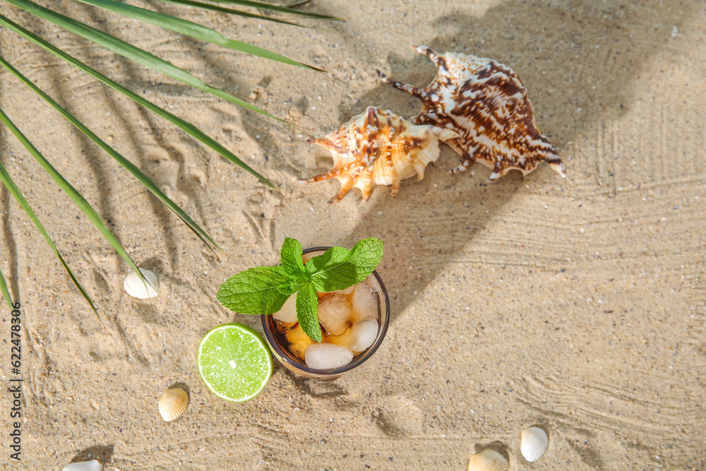 Glass of cold Cuba Libre cocktail and seashells on sand