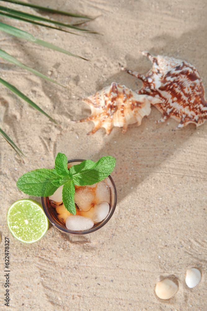 Glass of cold Cuba Libre cocktail and seashells on sand