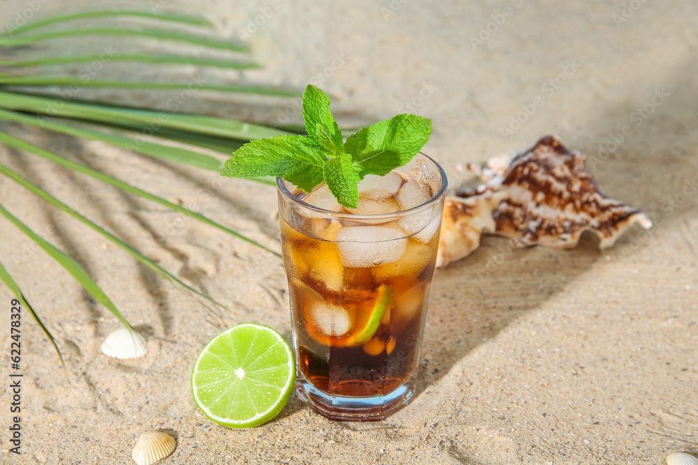 Glass of cold Cuba Libre cocktail and seashells on sand