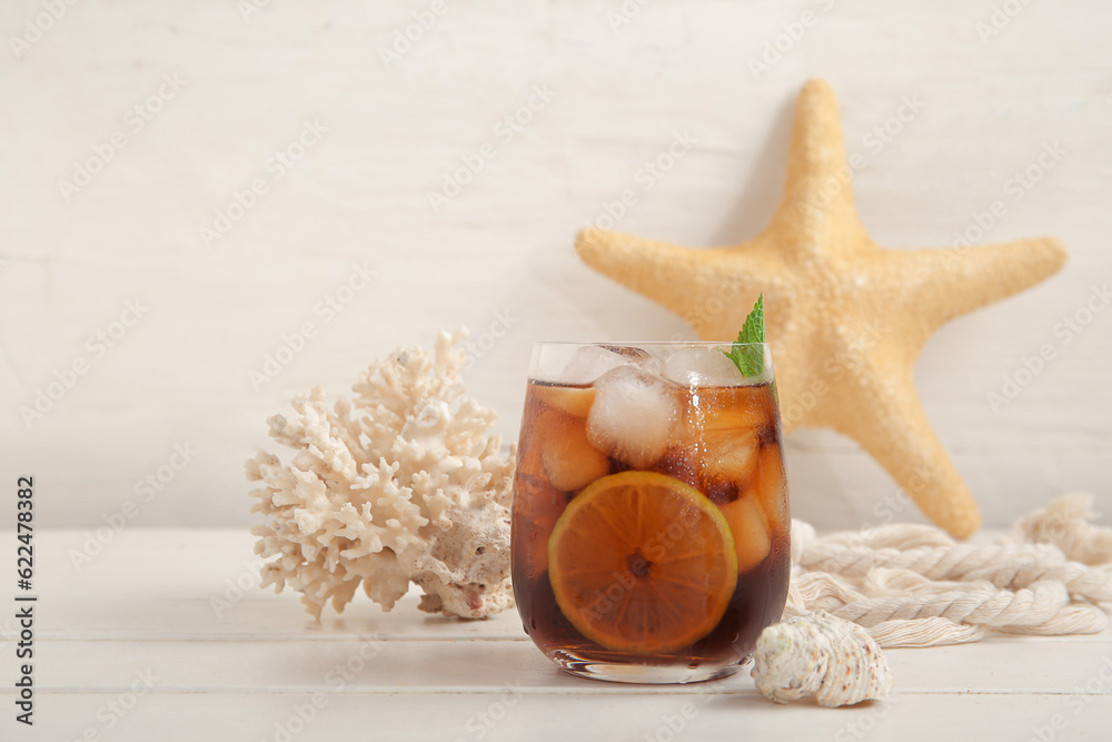 Glass of cold Cuba Libre cocktail, coral, seashell and starfish on white wooden table