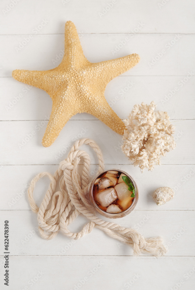 Glass of cold Cuba Libre cocktail, coral and starfish on white wooden background