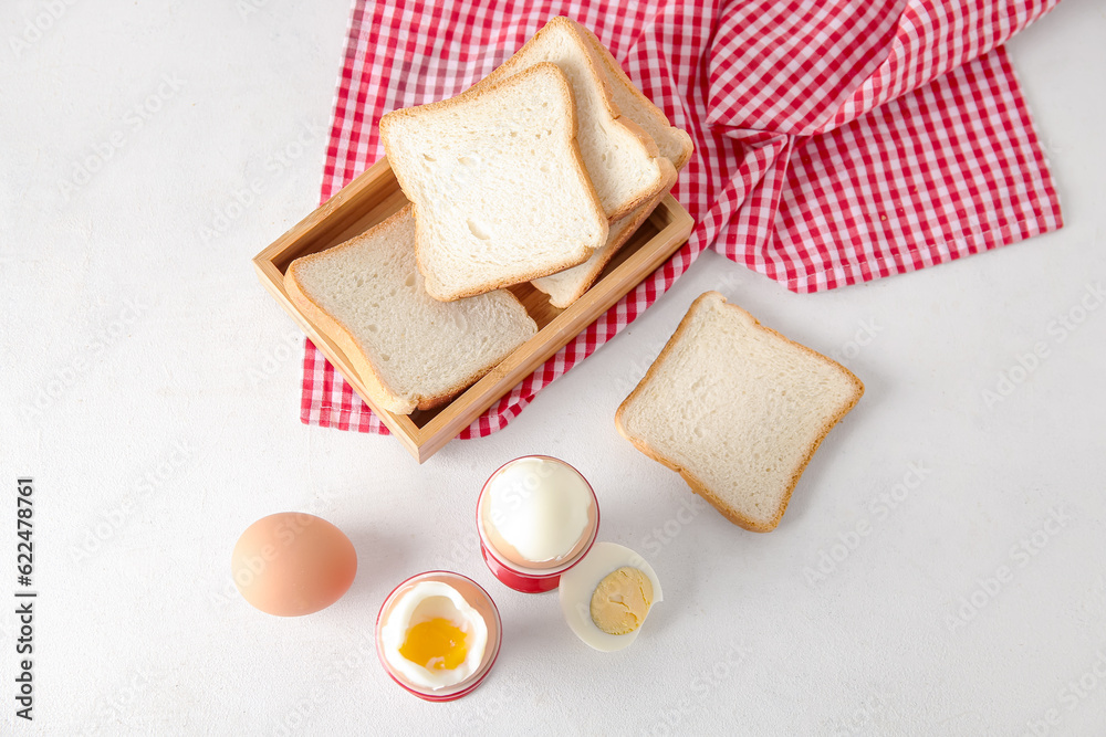 Holders with boiled chicken eggs and wooden box of bread on white background