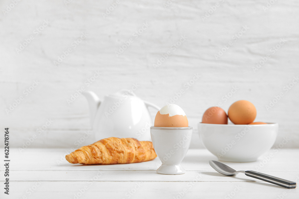 Holder with boiled chicken egg, teapot and croissant on white wooden background