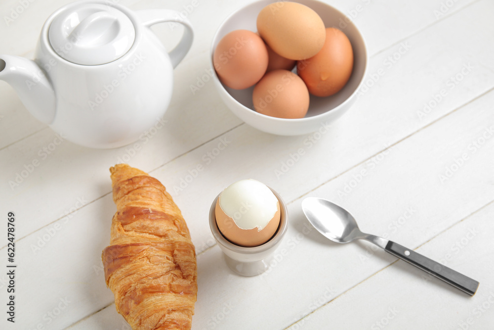 Holder with boiled chicken egg, teapot and croissant on white wooden background