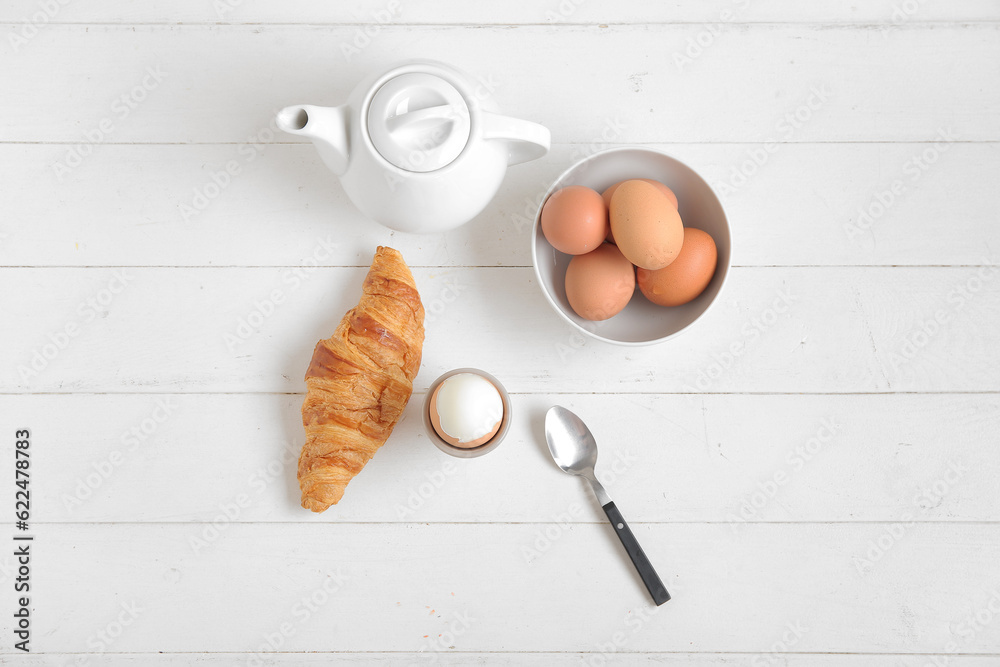 Holder with boiled chicken egg, teapot and croissant on white wooden background