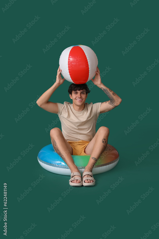 Young man with swim ring and beach ball on green background