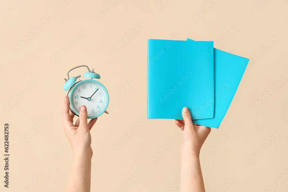 Female hands holding alarm clock and copybooks on beige background