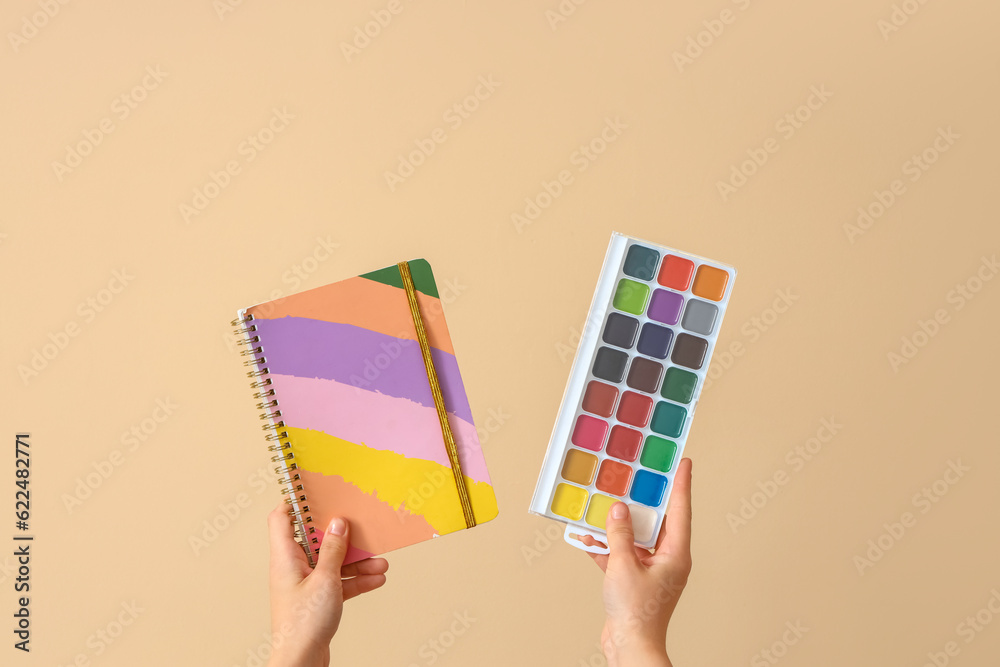 Female hands holding copybook and paints on beige background