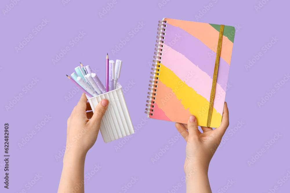 Female hands holding cup with pencils and copybook on lilac background