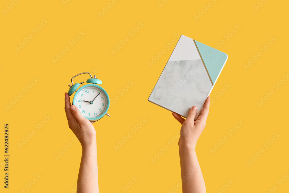 Female hands holding alarm clock and notebook on yellow background