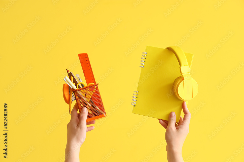 Female hands holding cup with stationery, notebook and headphones on yellow background