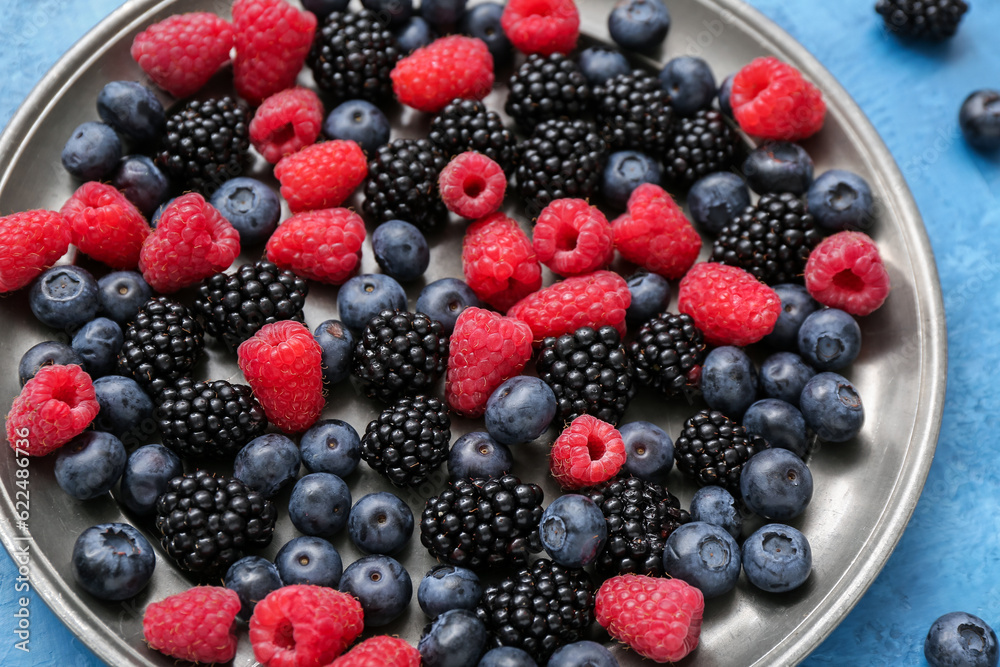 Plate with different fresh berries on blue background, closeup