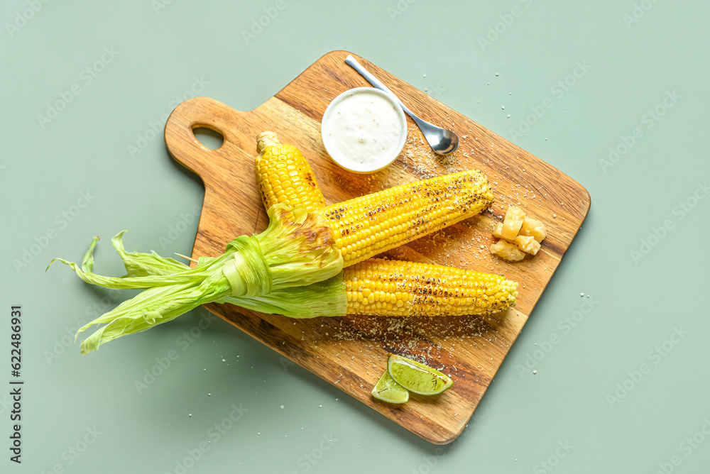 Wooden board with tasty grilled corn cobs and sauce on green background