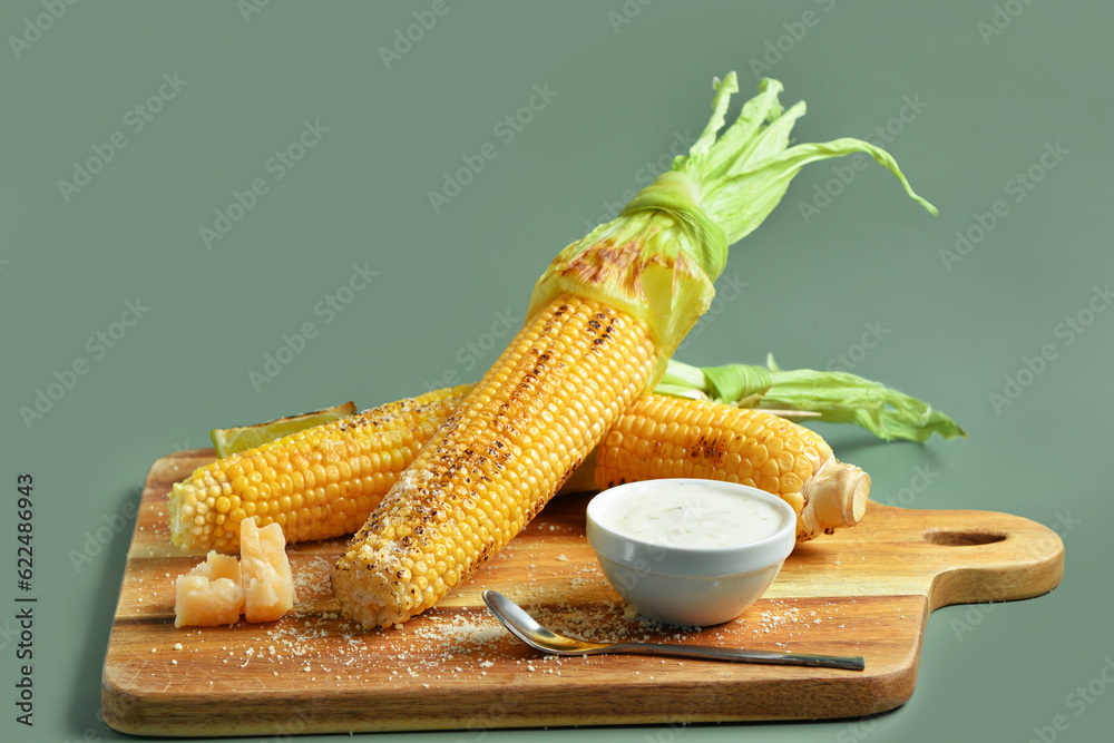 Wooden board with tasty grilled corn cobs and sauce on green background