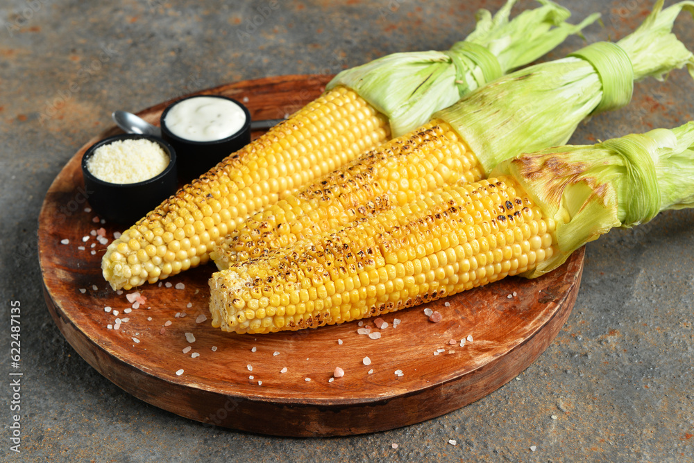 Wooden board with tasty grilled corn cobs and sauce on dark background