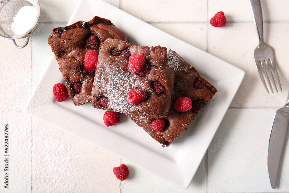 Plate with pieces of raspberry chocolate brownie on white tile background