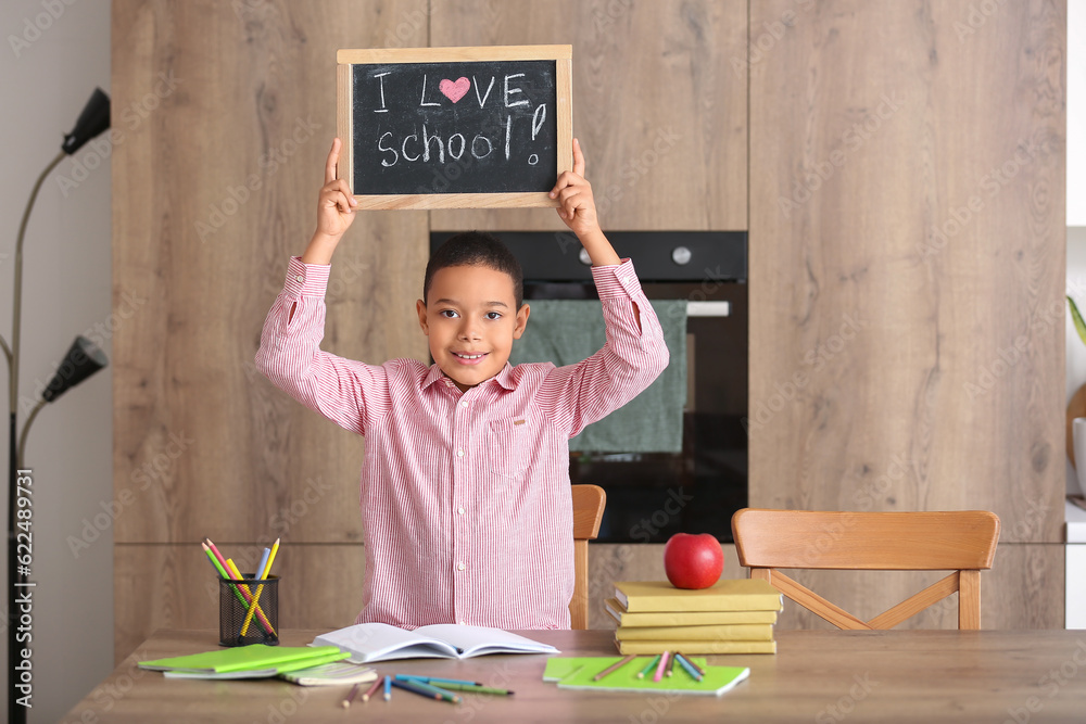 Little African-American boy holding chalkboard with text I LOVE SCHOOL in kitchen