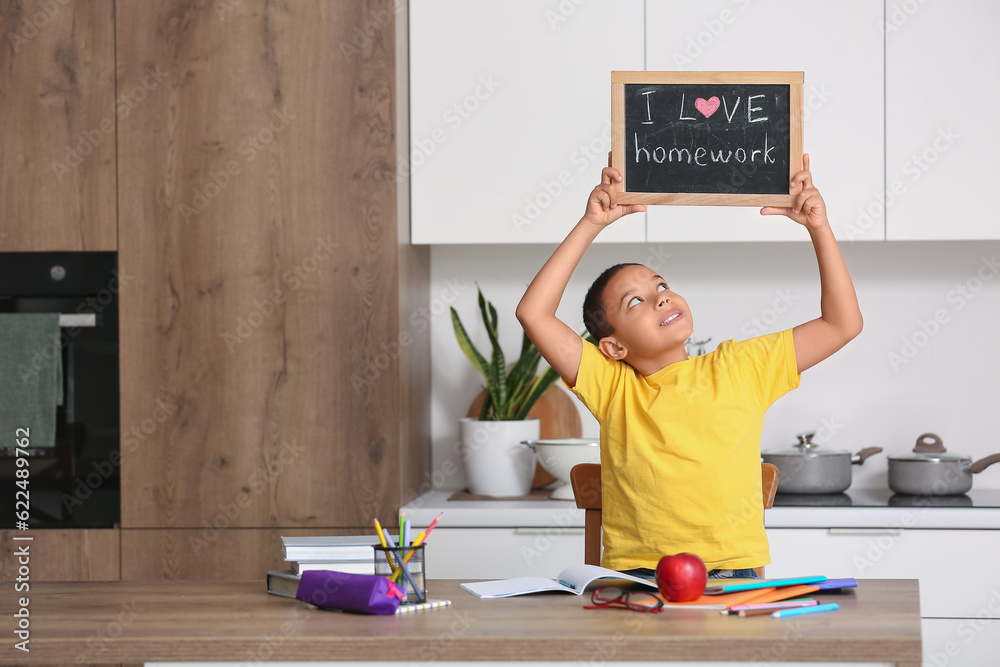 Little African-American boy holding chalkboard with text I LOVE HOMEWORK in kitchen