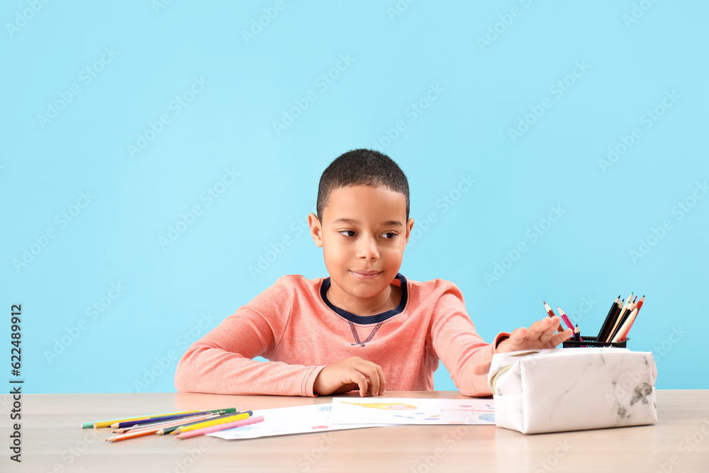 Little African-American boy with pencil case drawing at table on blue background