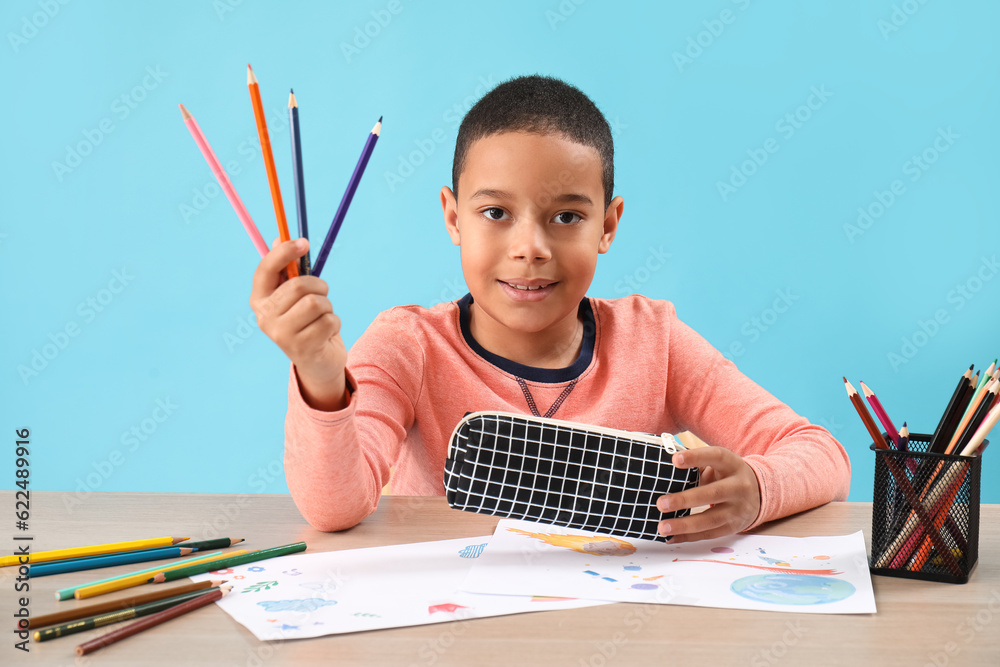 Little African-American boy with pencil case at table on blue background