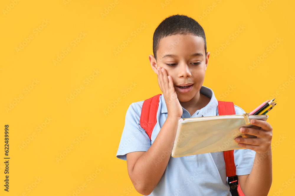 Surprised little African-American schoolboy with pencil case on orange background