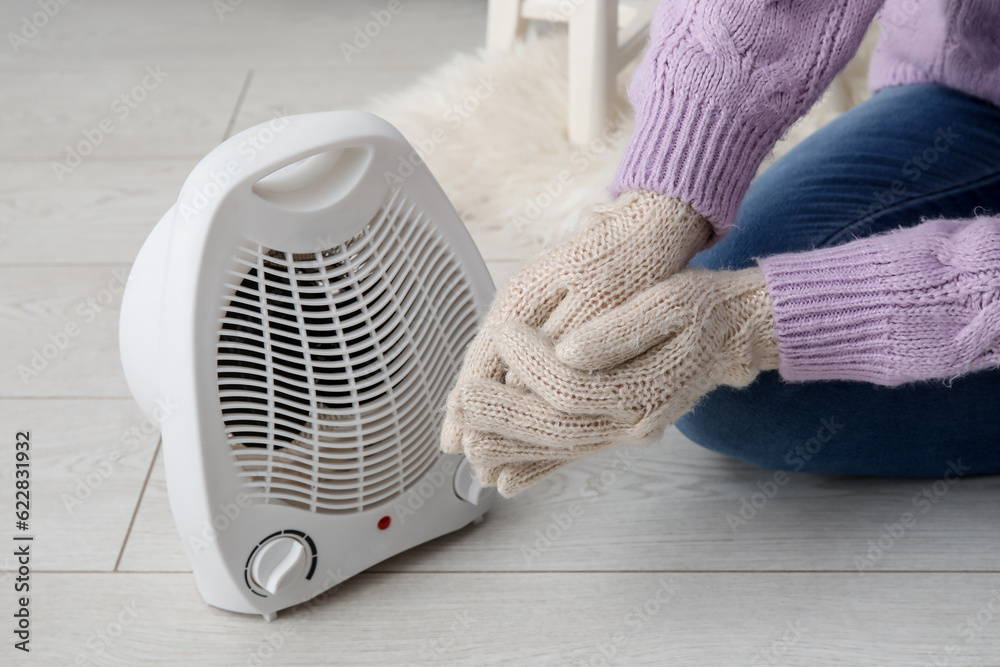 Woman warming hands in gloves near electric fan heater at home, closeup