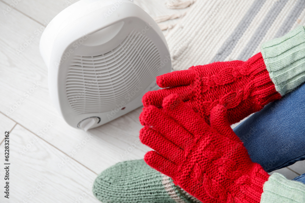 Woman warming hands in gloves near electric fan heater at home, closeup