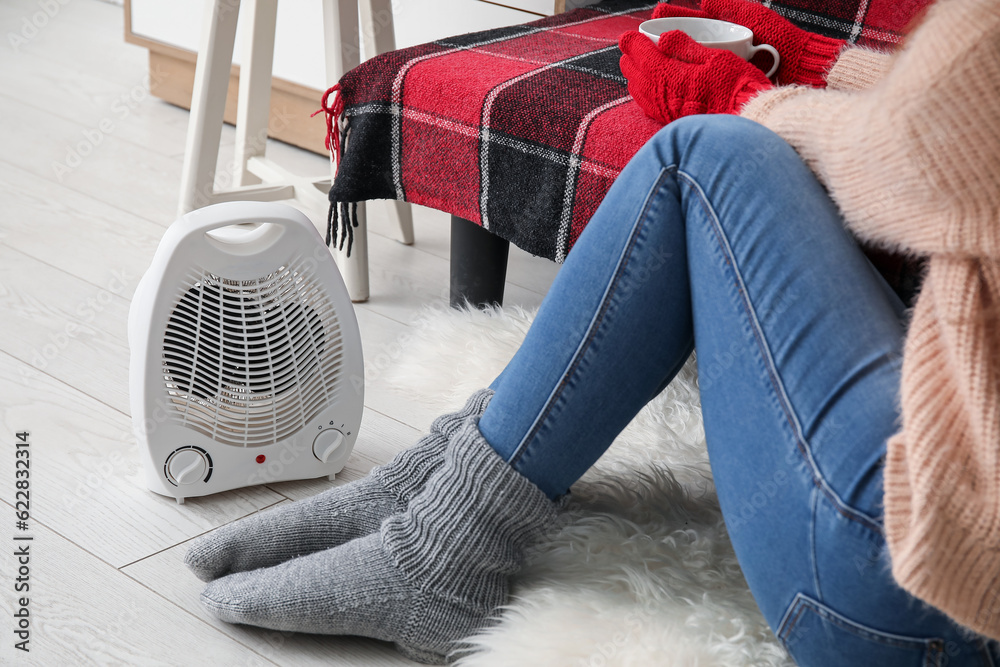 Woman warming legs in socks near electric fan heater at home, closeup