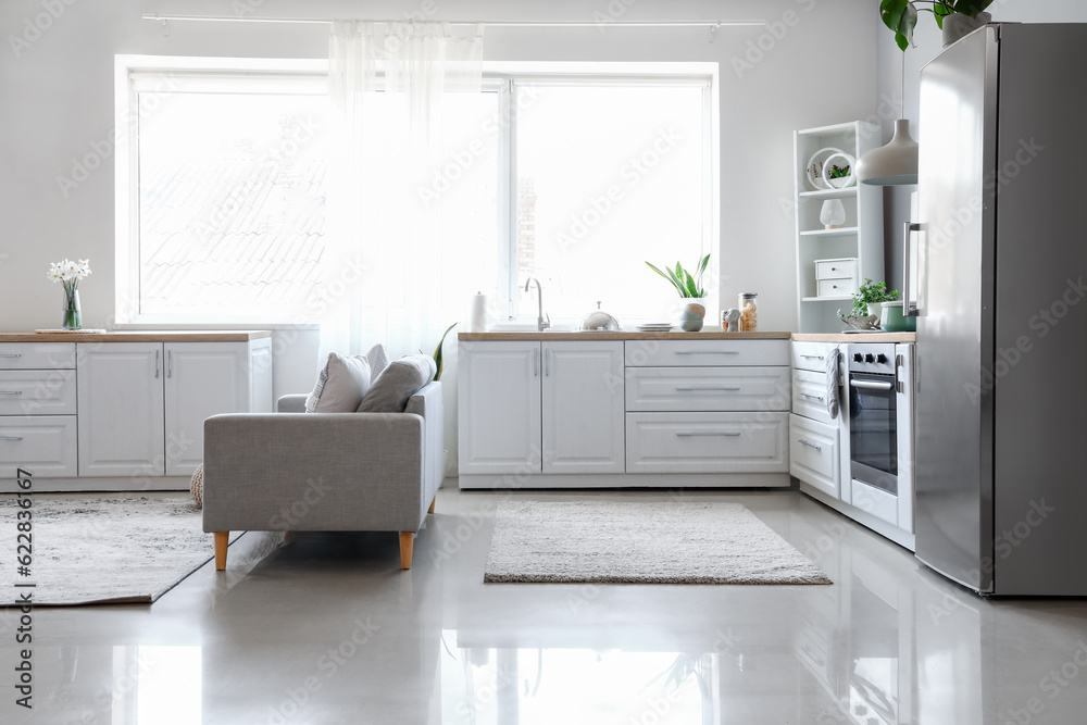 Interior of light kitchen with stylish fridge, counters, sofa and shelving unit