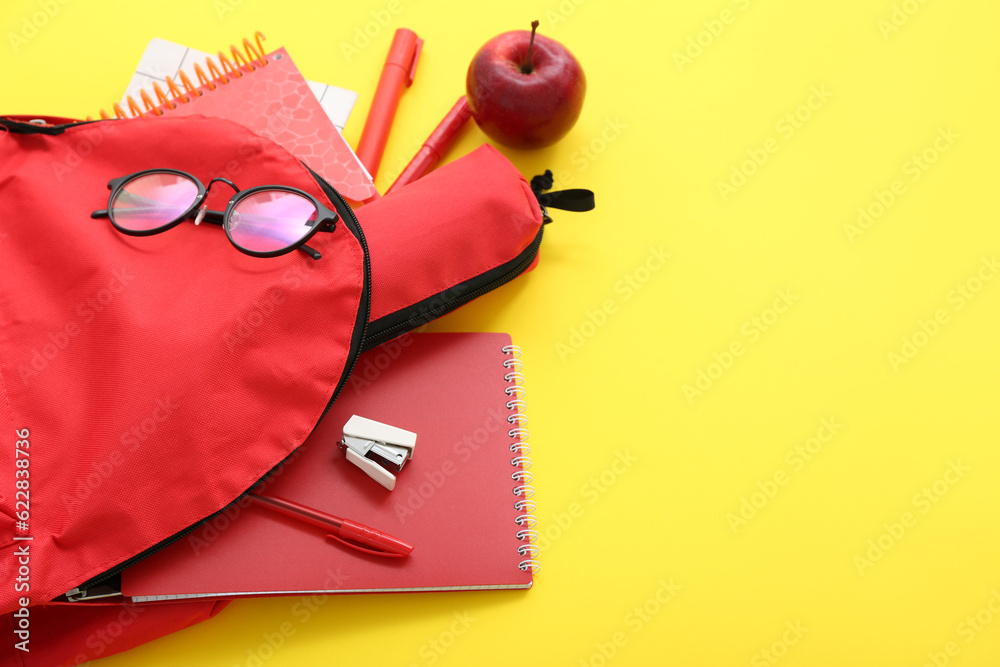 Red school backpack with notebooks, eyeglasses and pencil case on yellow background