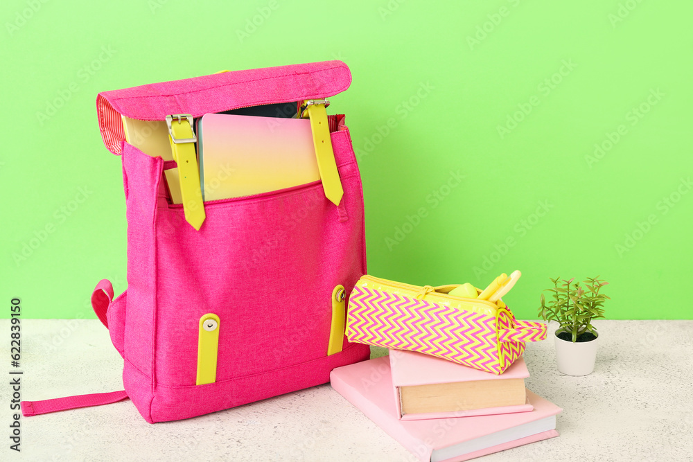 Pink school backpack with books, pencil case and houseplant on white wooden table near green wall