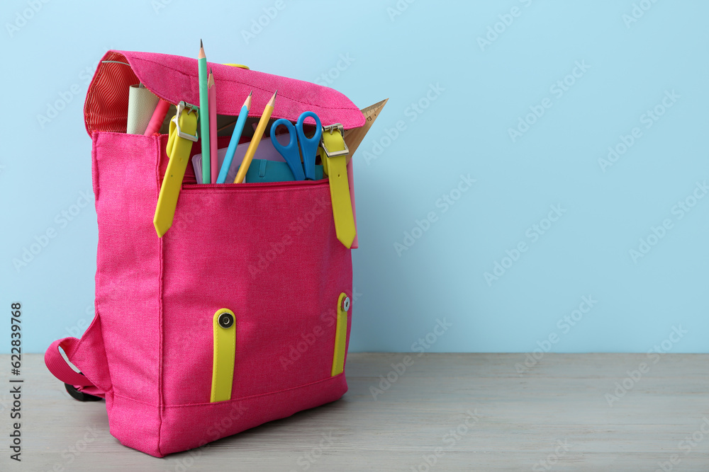 Pink school backpack with scissors, notebooks and pencils on grey wooden table near blue wall