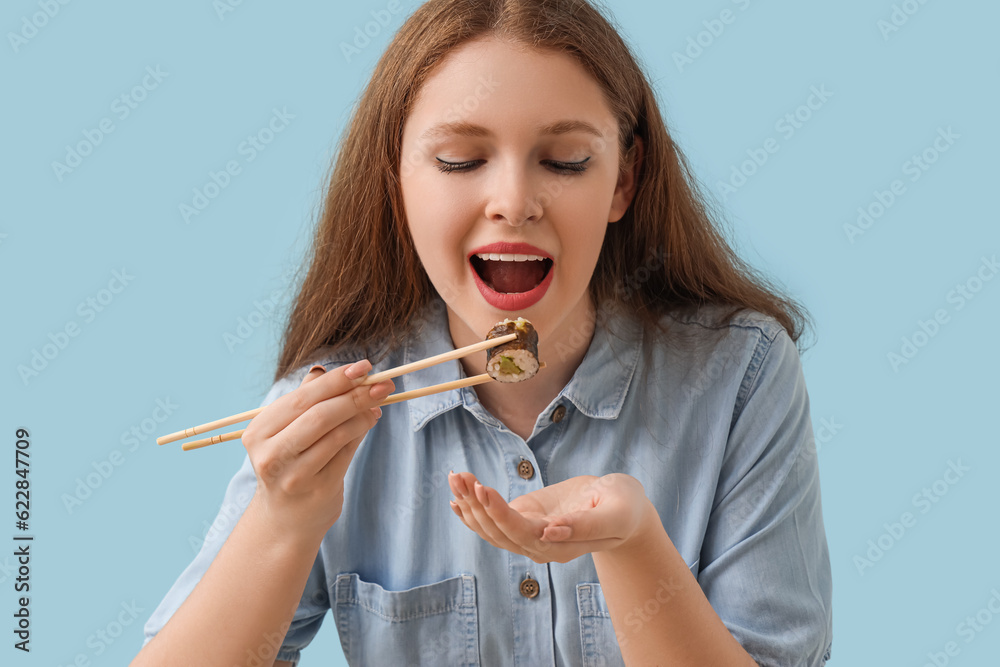 Young woman eating tasty sushi roll on blue background, closeup