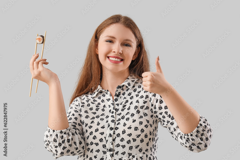 Young woman with tasty sushi roll showing thumb-up on grey background