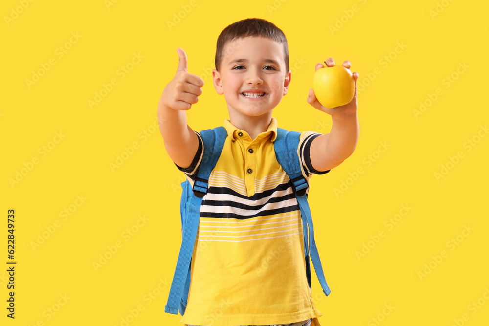 Little schoolboy with apple showing thumb-up on yellow background