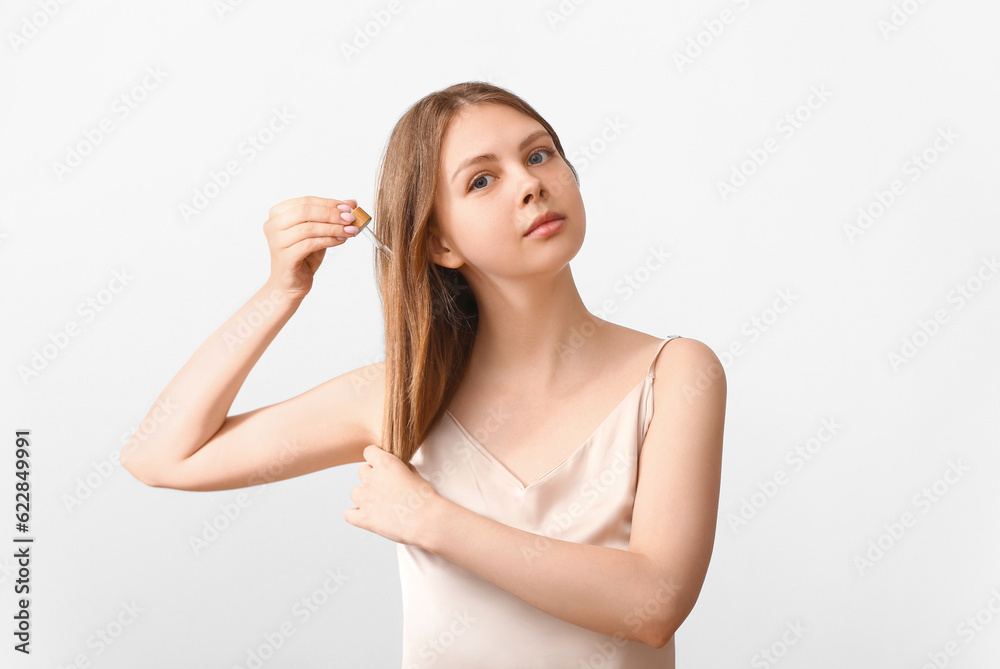 Young woman using cosmetic oil for hair treatment on light background