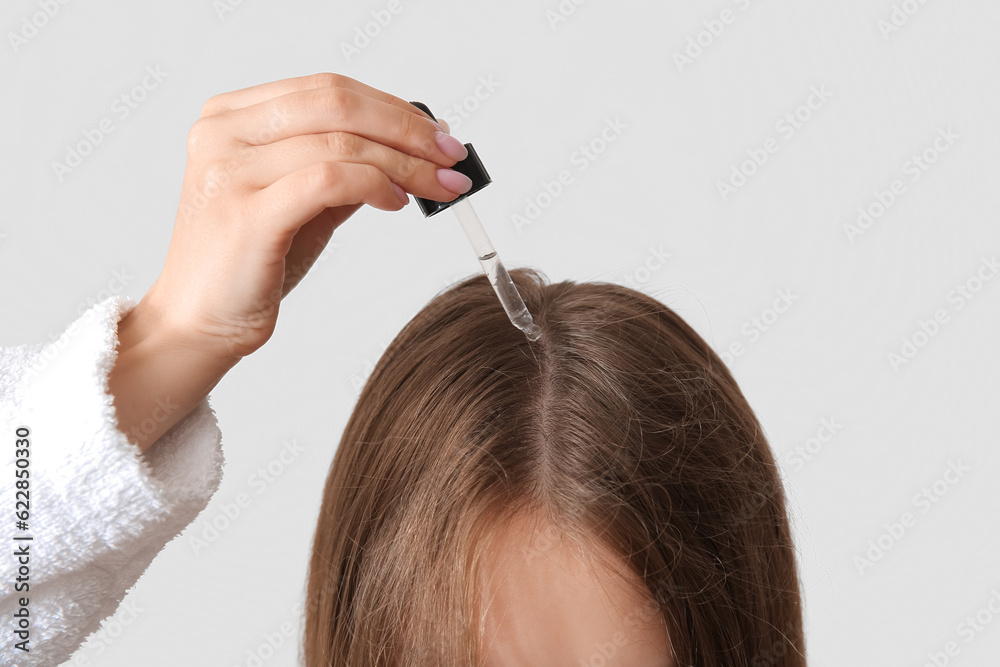 Young woman using cosmetic oil for hair treatment on light background, closeup