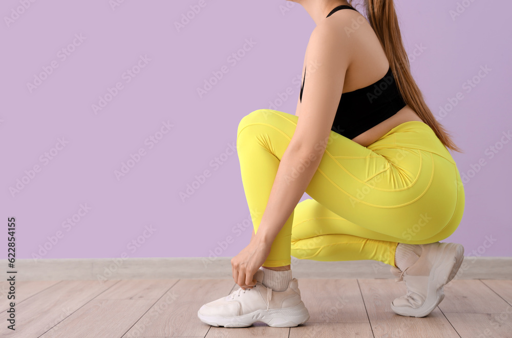 Young woman in sportswear tying shoelaces near lilac wall