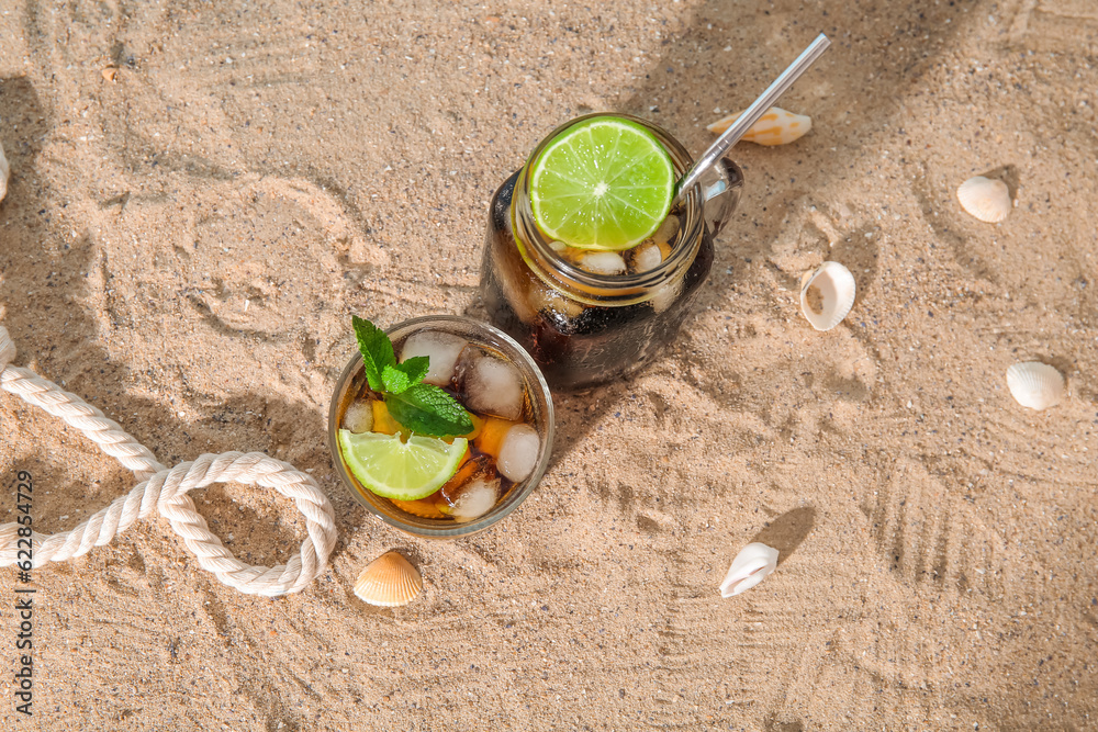 Glass and mason jar of cold Cuba Libre cocktail, seashells on sand