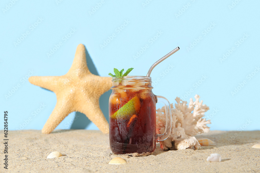 Mason jar of cold Cuba Libre cocktail, coral, starfish and seashells on sand near blue background