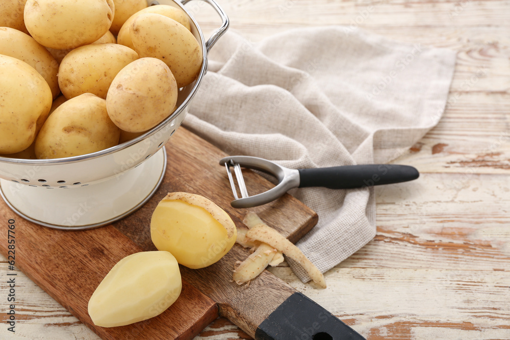 Colander and board with raw potatoes on white wooden background