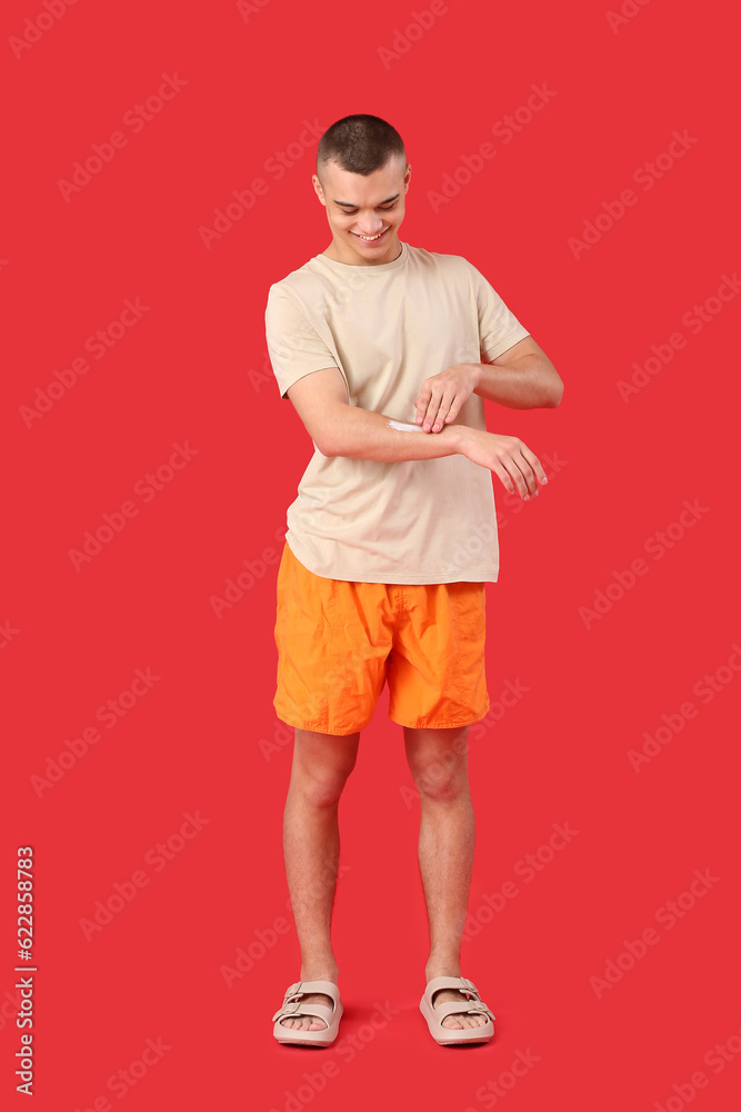 Happy young man applying sunscreen cream on his body against red background