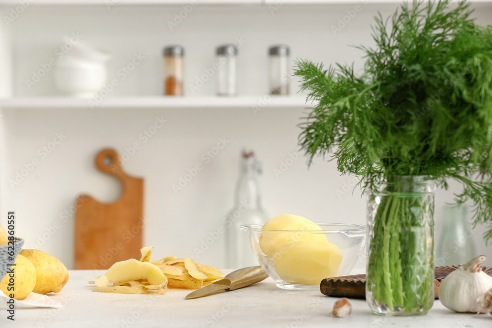 Bowl with peeled raw potatoes and dill on table in kitchen