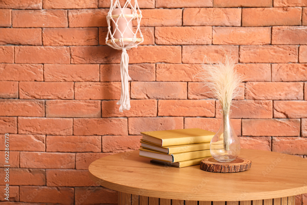 Vase with dried grass and books on wooden coffee table near brick wall