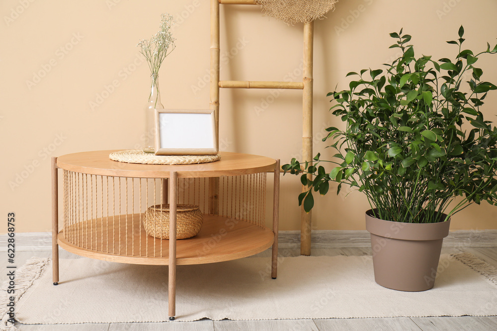 Wooden coffee table with blank frame and houseplant near beige wall